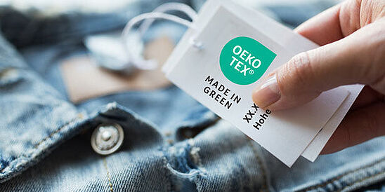 Chaqueta en mezclilla con mano sujetando una etiqueta OEKO-TEX® MADE IN GREEN