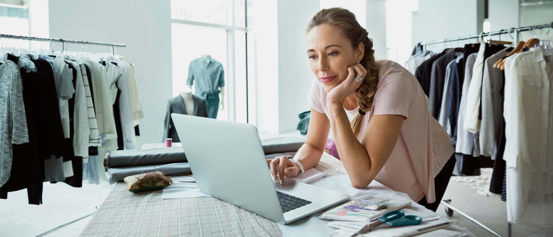 Diseñador en ropa en la computadora en la habitación con estantes en ropa